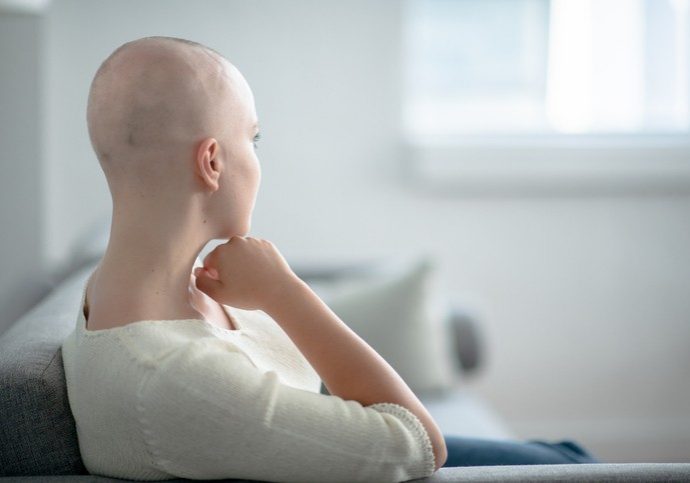 A young woman of Caucasian ethnicity is indoors at home. She appears to be thinking about her life. Her head is bald due to chemotheraphy.
