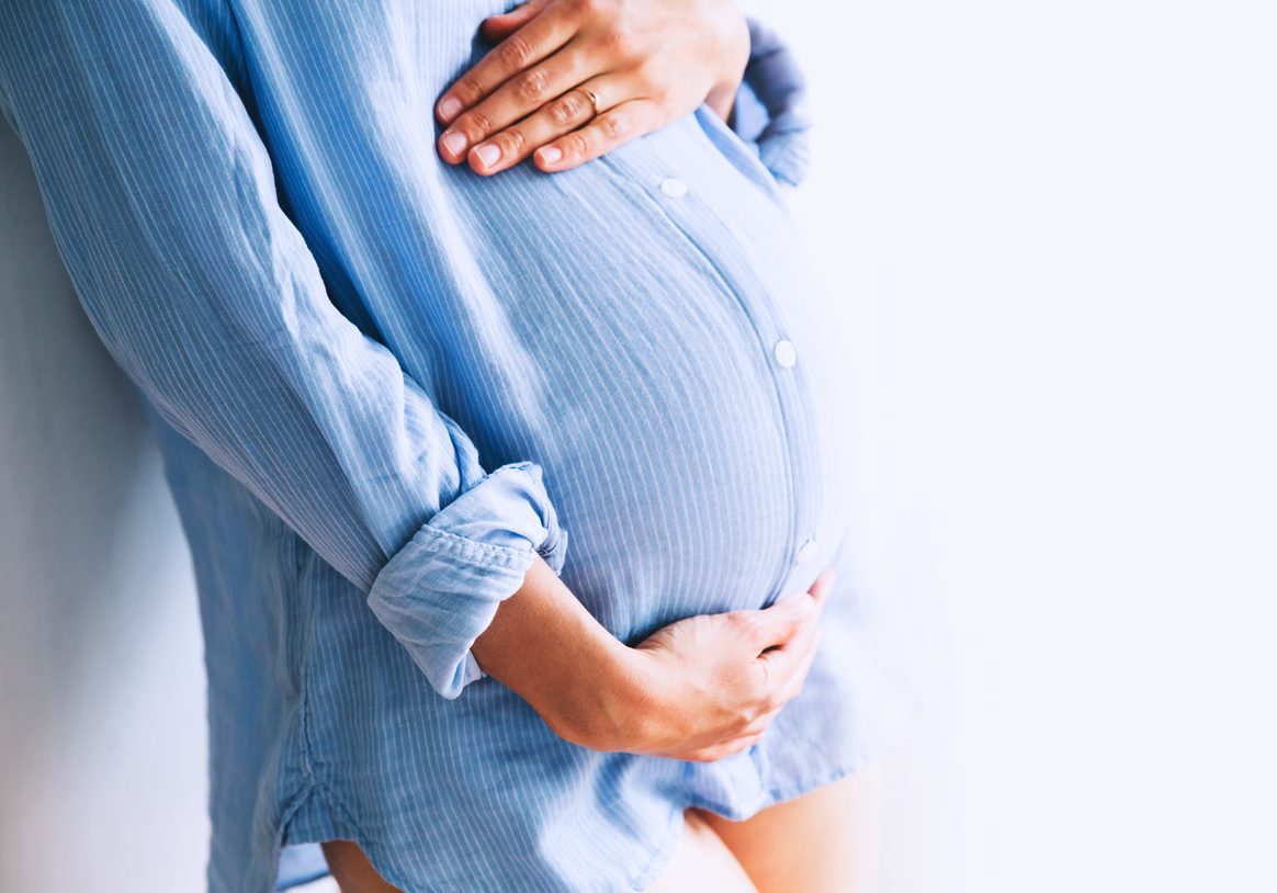 Pregnant woman holds hands on belly at home interiors. Pregnancy, parenthood, preparation and expectation concept. Close-up, copy space, indoors.